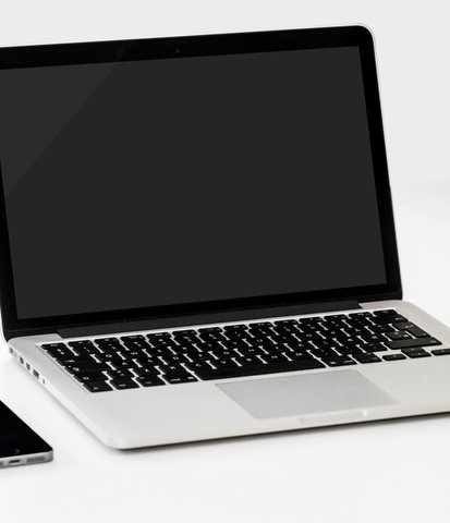 A laptop with a blank screen sits on a white desk, flanked by a smartphone and camera lens cap on the left and a red apple on the right.